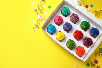 Photo of Box with different cupcakes and confetti on yellow background, flat lay. Space for text