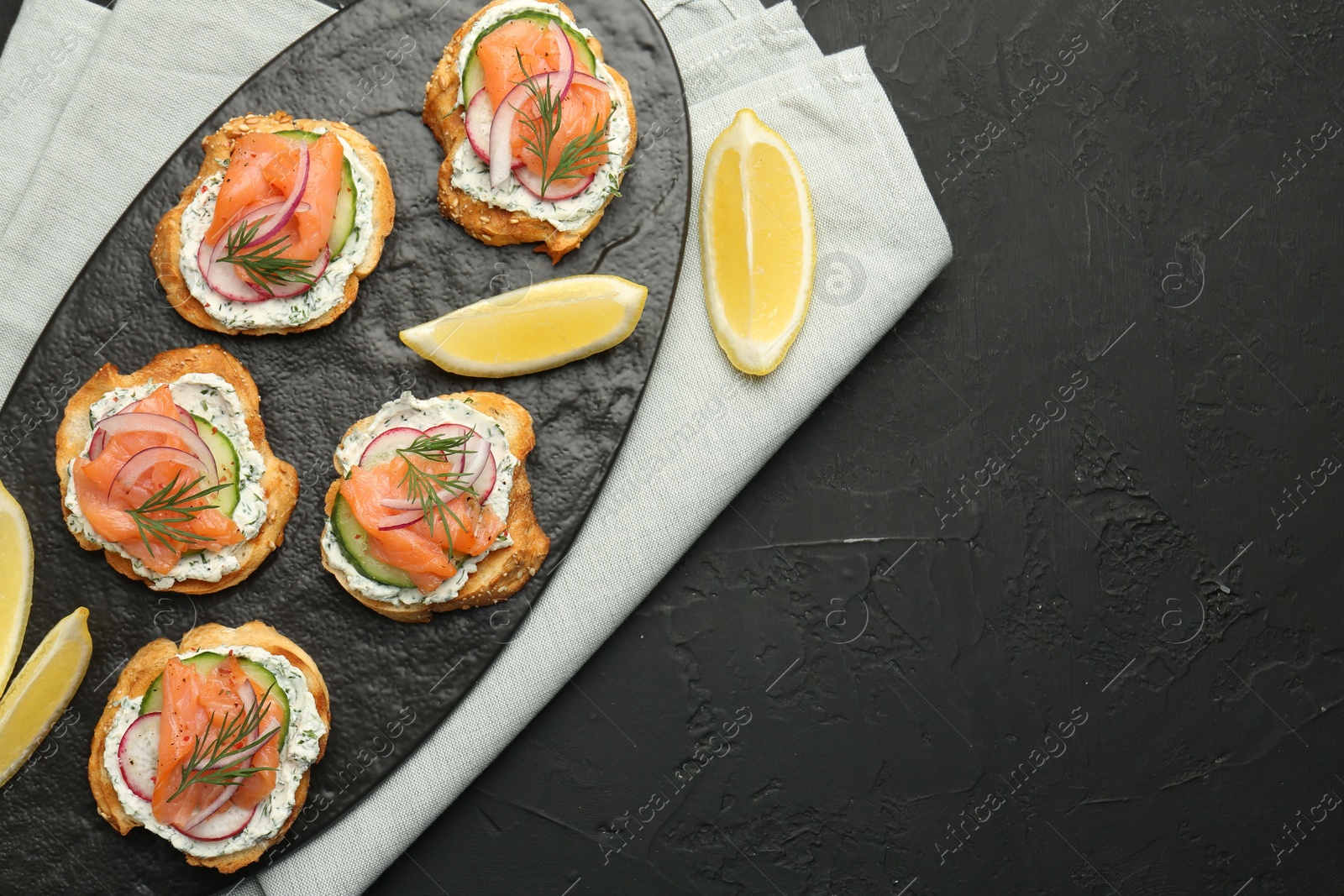 Photo of Tasty canapes with salmon, cucumber, radish and cream cheese on black table, top view. Space for text