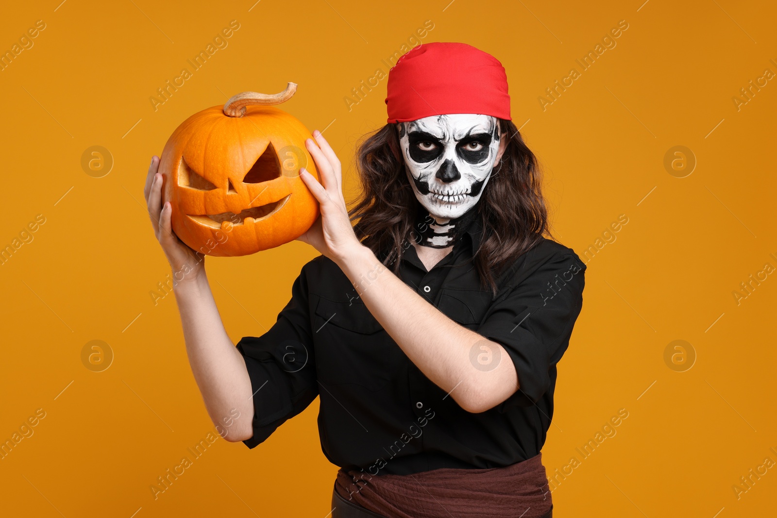 Photo of Man in scary pirate costume with skull makeup and carved pumpkin on orange background. Halloween celebration