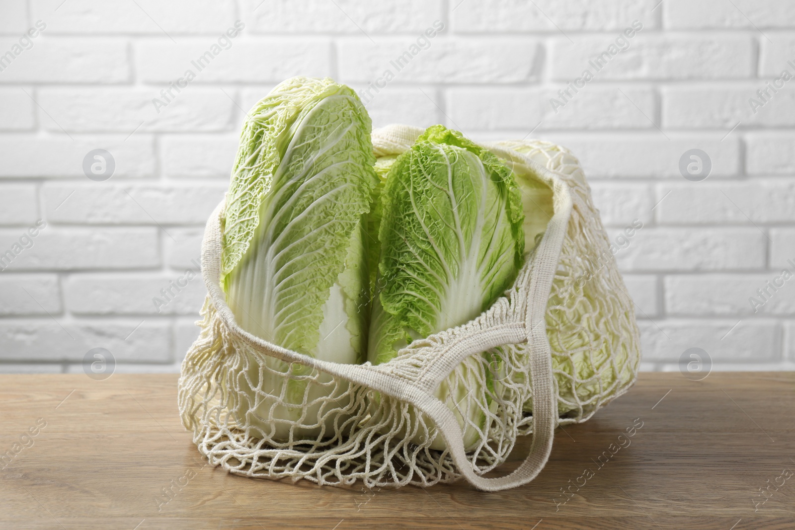 Photo of Fresh Chinese cabbages in string bag on wooden table near white brick wall