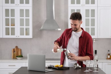 Man learning to make cocktail with online video on laptop at table in kitchen. Time for hobby