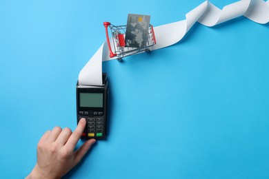 Photo of Man using payment terminal with credit card, small shopping cart and thermal paper for receipt on light blue background, top view