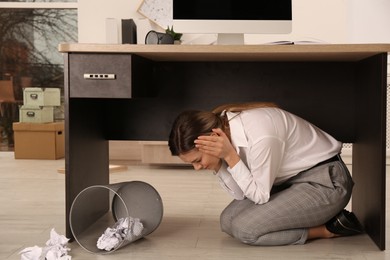 Scared young woman hiding under office desk during earthquake