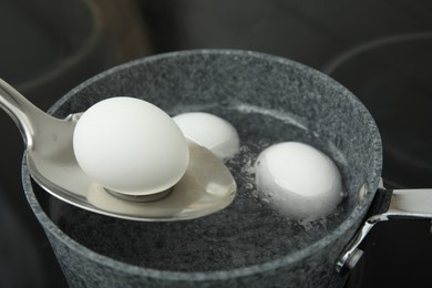 Spoon with boiled egg above saucepan on electric stove, closeup