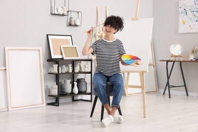 Photo of Young woman with brush and artist`s palette near easel in studio