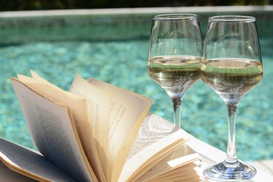 Photo of Glasses of tasty wine and open book on wooden table near swimming pool, closeup