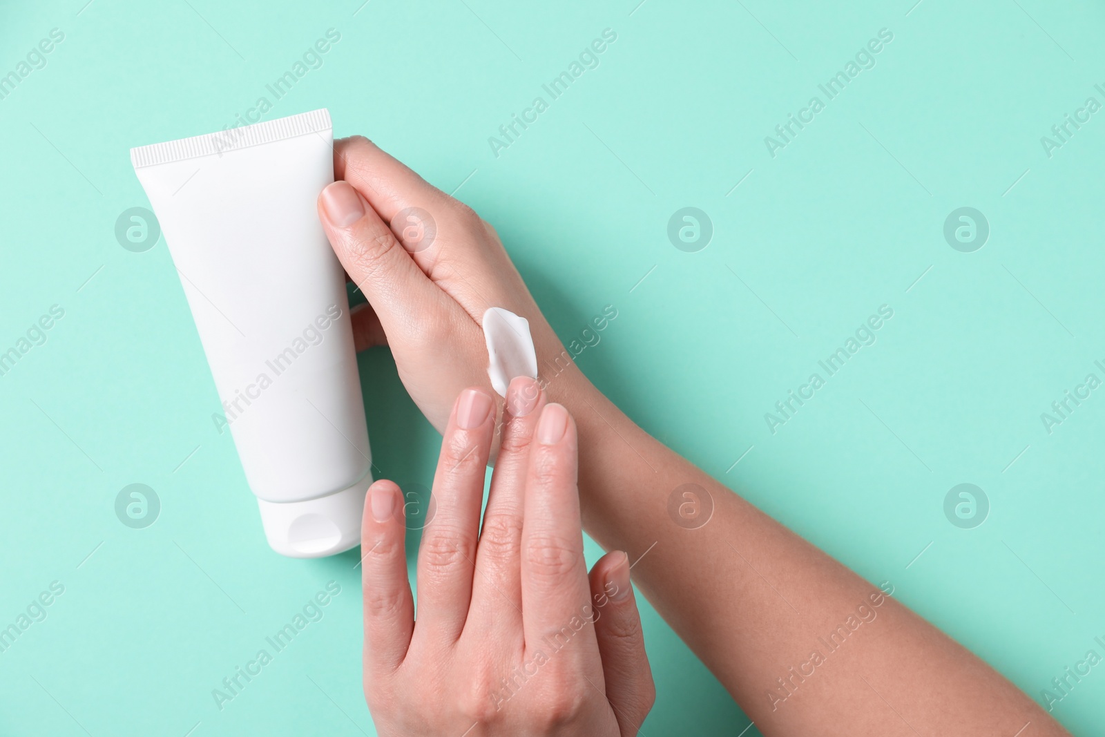 Photo of Woman with tube applying cosmetic cream onto her hand on turquoise background, top view