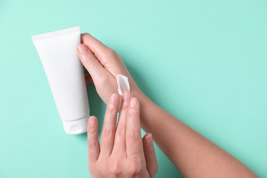 Woman with tube applying cosmetic cream onto her hand on turquoise background, top view