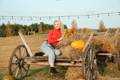 Photo of Beautiful woman with bouquet sitting on wooden cart with pumpkins and hay in field. Autumn season