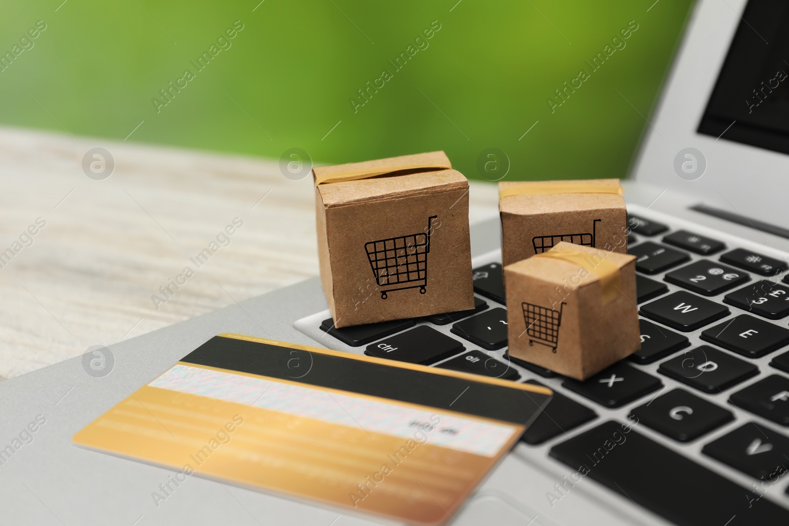 Image of Internet store. Small cardboard boxes with shopping carts, credit card and laptop on light wooden table, closeup