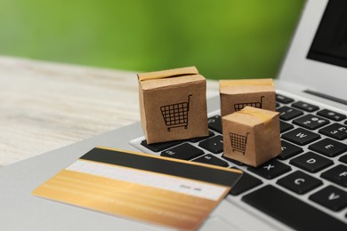 Image of Internet store. Small cardboard boxes with shopping carts, credit card and laptop on light wooden table, closeup