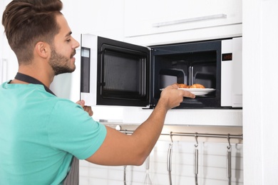 Photo of Young man putting plate with croissants into microwave oven at home