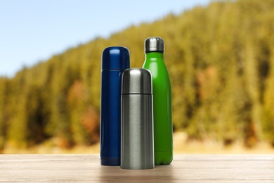 Image of Different thermos bottles on wooden table against blurred mountain