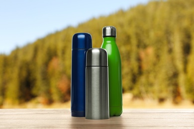 Different thermos bottles on wooden table against blurred mountain