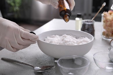 Photo of Woman in gloves adding essential oil to bath bomb mixture at grey table, closeup