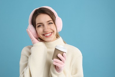 Photo of Happy woman with cup of drink wearing warm earmuffs on light blue background