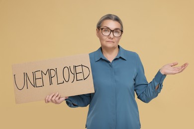 Photo of Confused senior woman holding cardboard sign with word Unemployed on beige background