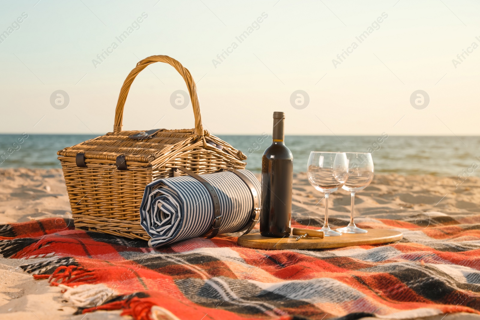 Photo of Blanket with picnic basket, corkscrew, bottle of wine and glasses on sandy beach near sea