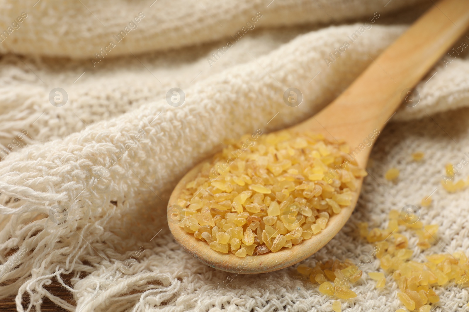 Photo of Wooden spoon with raw bulgur on beige cloth, closeup