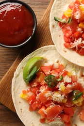 Delicious tacos with vegetables, lime and ketchup on wooden table, flat lay