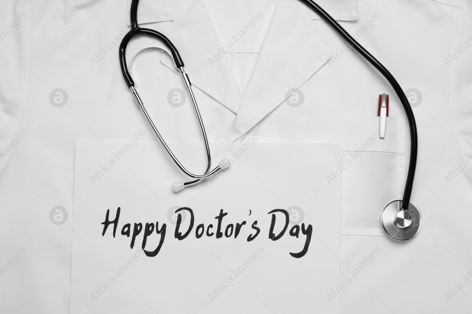 Photo of Stethoscope and paper sheet with phrase Happy Doctor's Day on white medical uniform, top view