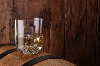 Whiskey with ice cubes in glass on barrel against wooden background, space for text