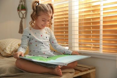 Photo of Cute little girl reading book near window at home