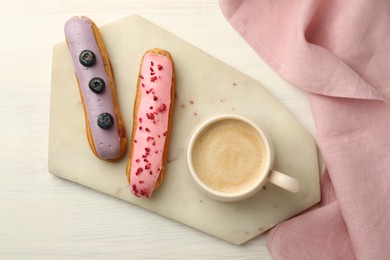 Cup of coffee and delicious eclairs covered with glaze on white wooden table, top view