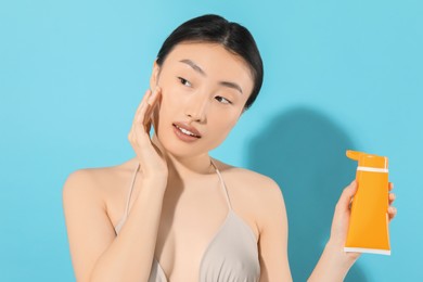 Beautiful young woman holding sun protection cream on light blue background