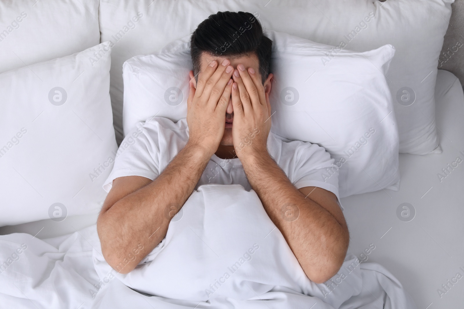 Photo of Man covering his face with hands while lying in bed at home, top view