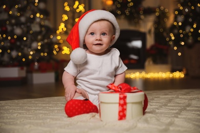 Photo of Cute baby in Santa hat with Christmas gift on floor at home