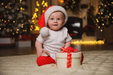 Cute baby in Santa hat with Christmas gift on floor at home