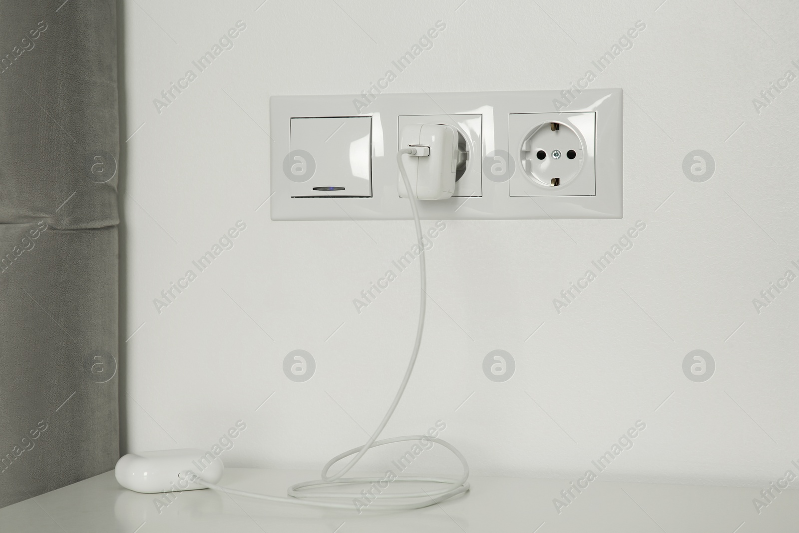 Photo of Charging case of modern wireless earphones plugged into power socket on white table indoors