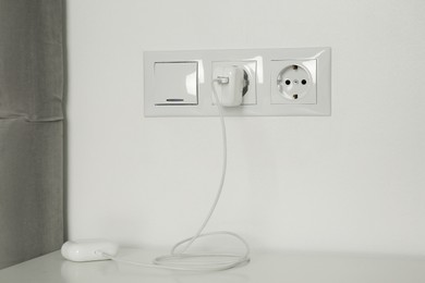 Photo of Charging case of modern wireless earphones plugged into power socket on white table indoors