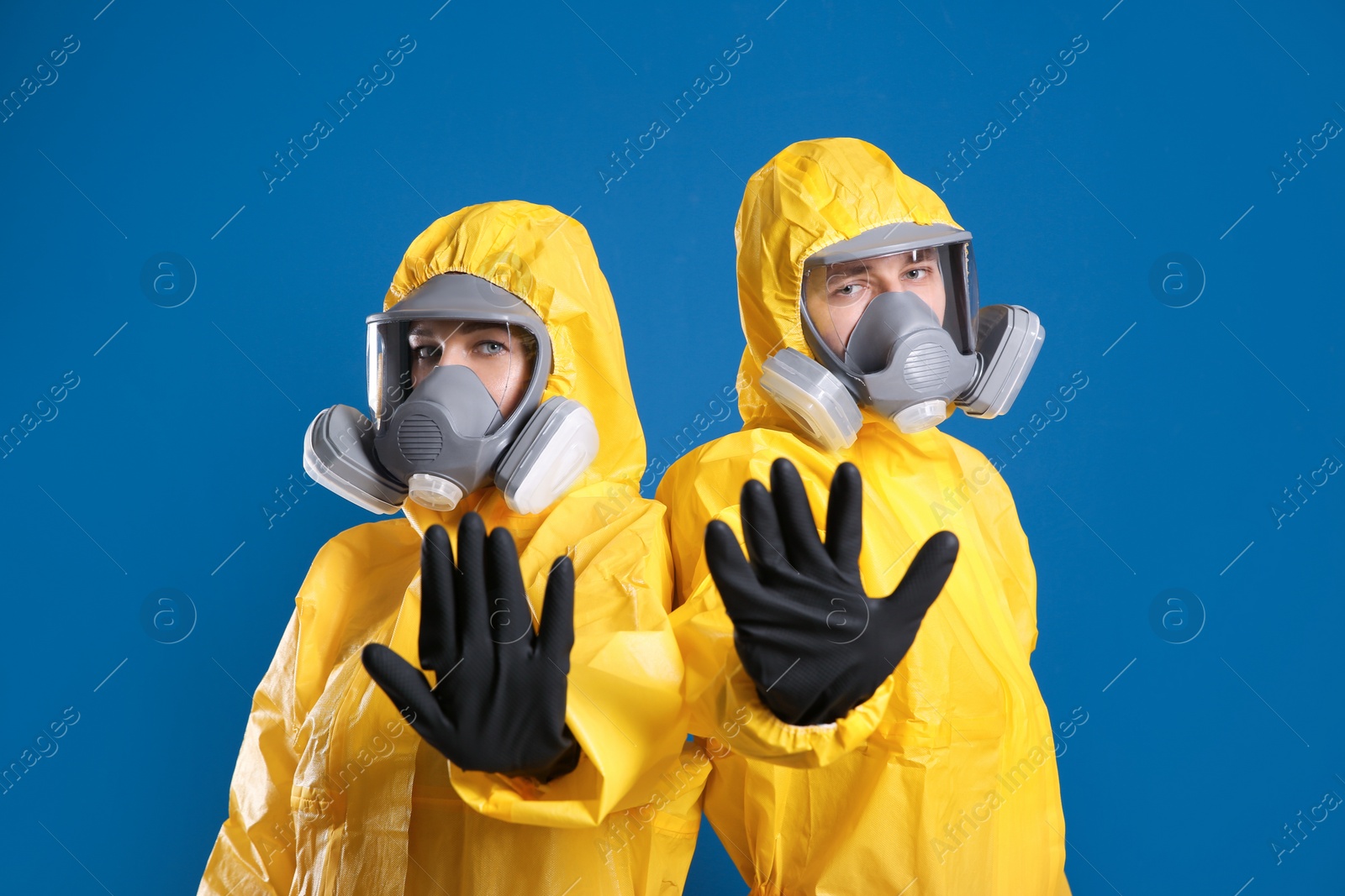 Photo of Man and woman in chemical protective suits making stop gesture on blue background. Virus research