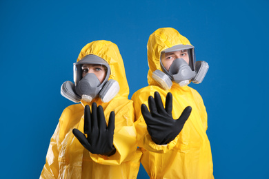 Photo of Man and woman in chemical protective suits making stop gesture on blue background. Virus research