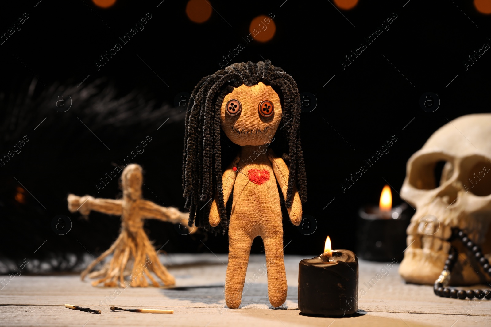 Photo of Female voodoo doll with pin in heart and ceremonial items on wooden table against blurred background