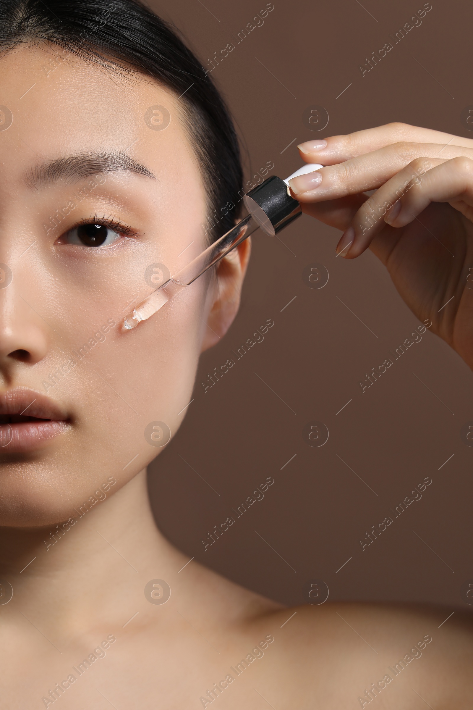 Photo of Beautiful young woman applying cosmetic serum onto her face on brown background, closeup