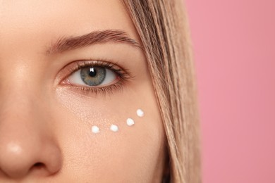 Photo of Young woman with cream around eye on pink background, closeup. Space for text