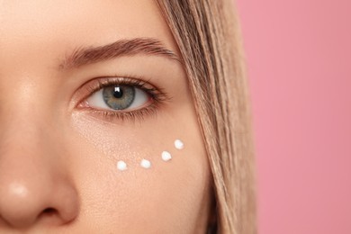 Young woman with cream around eye on pink background, closeup. Space for text