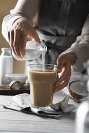 Woman pouring coconut milk into glass of coffee at white wooden table, closeup