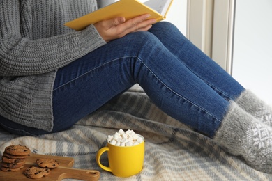 Photo of Woman with cup of hot cocoa reading book on window sill, closeup. Winter drink