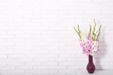 Photo of Vase with beautiful gladiolus flowers on table against white brick wall. Space for text