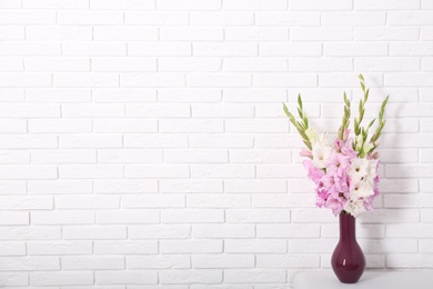 Vase with beautiful gladiolus flowers on table against white brick wall. Space for text