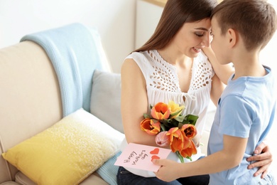 Happy woman receiving flowers and greeting card from her son at home. Mother's day celebration