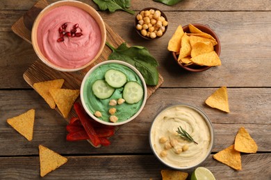 Photo of Different kinds of tasty hummus served with nachos on wooden table, flat lay