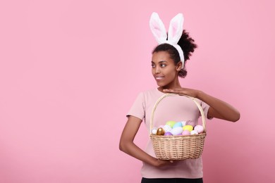Happy African American woman in bunny ears headband holding wicker basket with Easter eggs on pink background, space for text