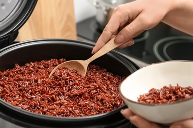 Photo of Woman putting brown rice into bowl from multi cooker in kitchen, closeup