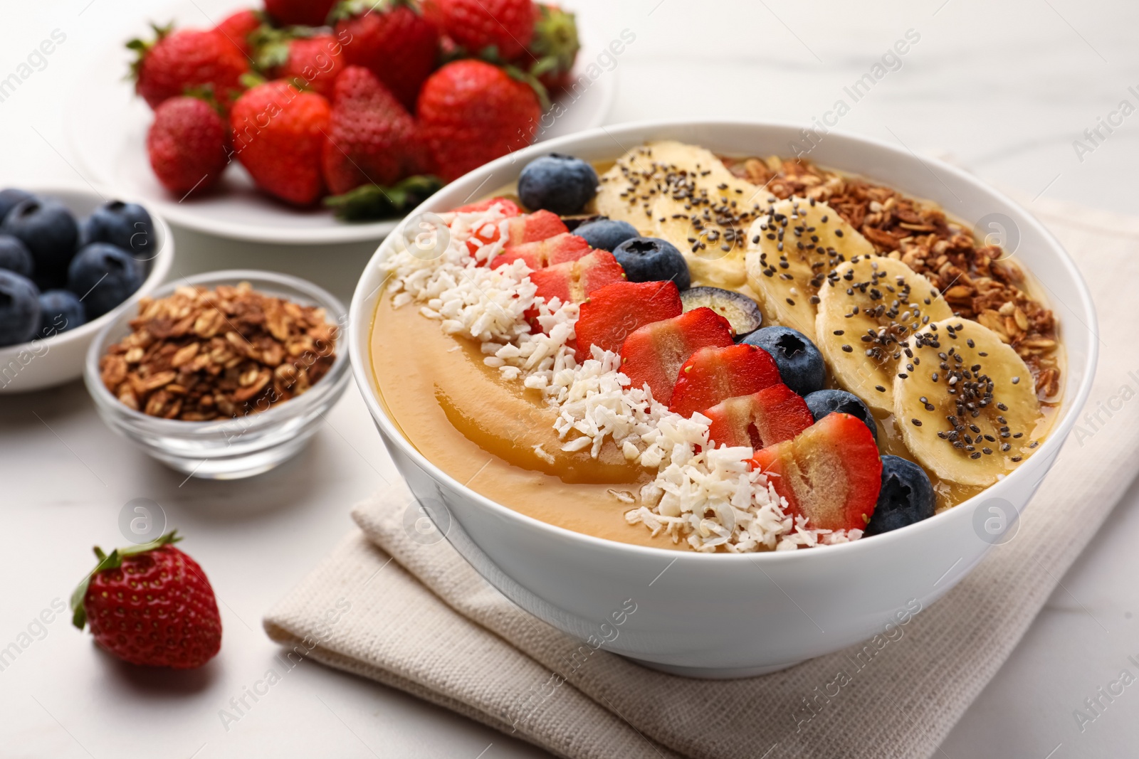 Photo of Delicious smoothie bowl with fresh berries, banana, coconut flakes and granola on white table, closeup
