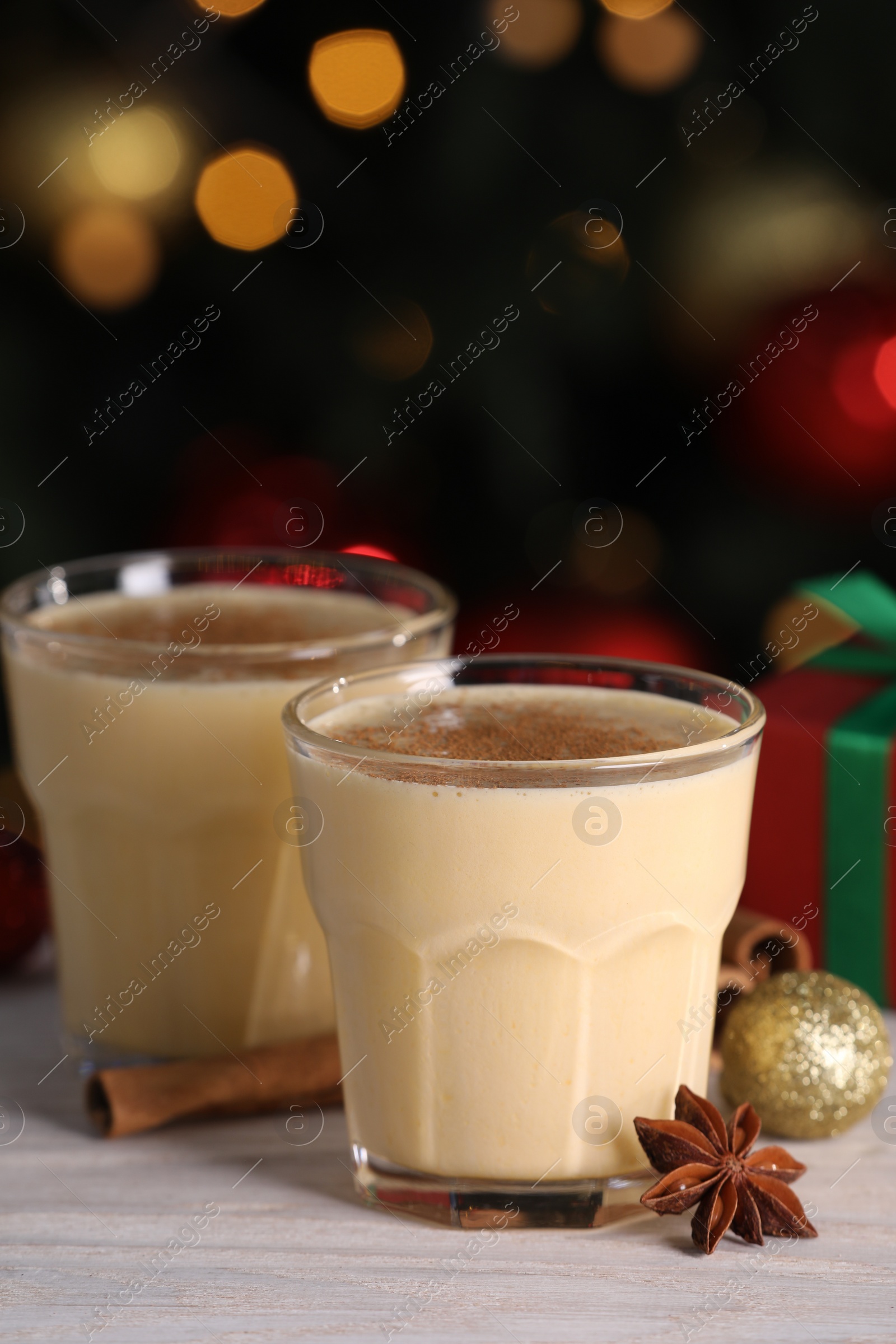 Photo of Tasty eggnog, cinnamon, anise and Christmas decorations on white wooden table against blurred lights